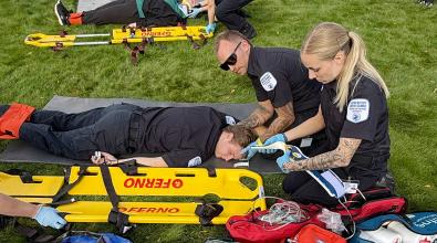 A male paramedic student with hands under the neck of another male paramedic student lying face down on a stretcher on the grass while a female paramedic student works with a bandage.