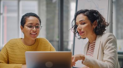A woman gestures to a laptop screen while another woman looks on.