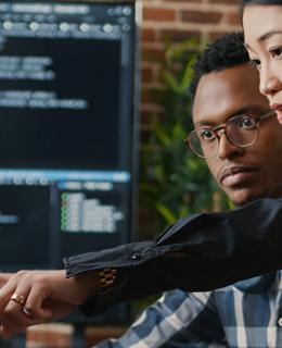Standing woman points at computer monitor while seated man looks on.