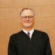 Man against beige wall wearing glasses and black graduation robe