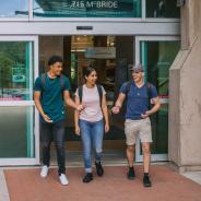 One female student walks out of campus doors flanked by two male students, all smiling in conversation.