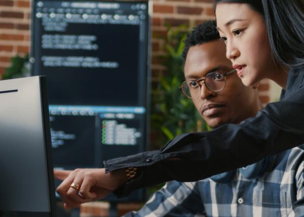 Standing woman points at computer monitor while seated man looks on.