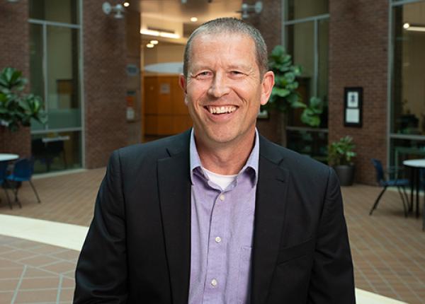 Man with short cropped hair wearing dark jacket and blue buttoned shirt.