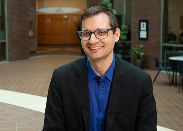 Dark haired man with glasses wearing black jacket and royal blue shirt.