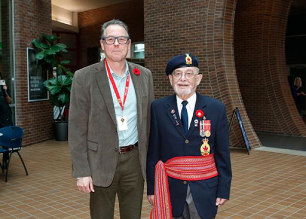 Man stands next to older man wearing military uniform with red Metis sash tied around his waist..