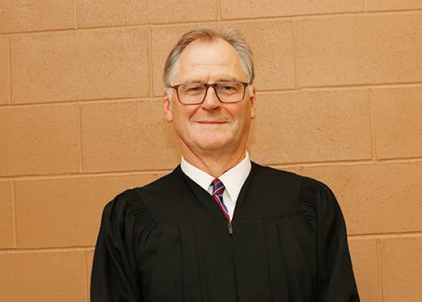 Man against beige wall wearing glasses and black graduation robe