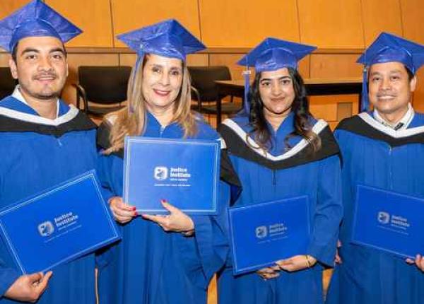 Four graduates in blue caps and gowns stand in front of wood-paneled walls, smiling and holding blue diploma folders with JIBC logo.